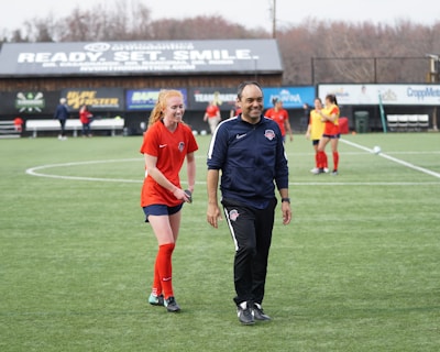 A group of people are on a soccer field, with some wearing sports attire. In the foreground, a person in red sports clothing is walking alongside someone in a dark tracksuit. Other people are visible in the background, some wearing yellow jerseys. A building with advertising banners, including a prominent sign reading 'Ready, Set, Smile,' is in the background.